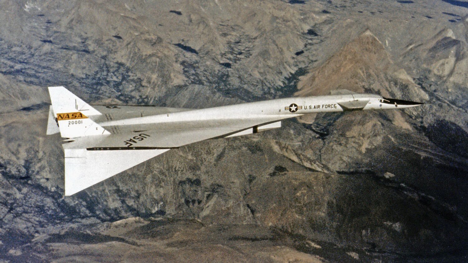 White delta-wing aircraft overflying mountains. The front of the fuselage features canard wings, and the wing tips are dropped.